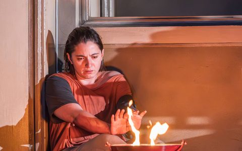 Pearl Chanda sits on the floor with a fire in a bucket in front of her, she sits with her hands outstretched towards the fire.