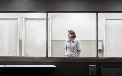 Pearl Chanda stands visible through a large window. She wears a camel coloured dress and her hands are in her pocket.