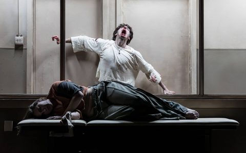 Pearl Chanda lies sideways on a cream doctor's bed. Behind her, visible through a window, Luke Cinque-White stands with his arms outstretched, his mouth is open and covered in blood and his head is tipped back.