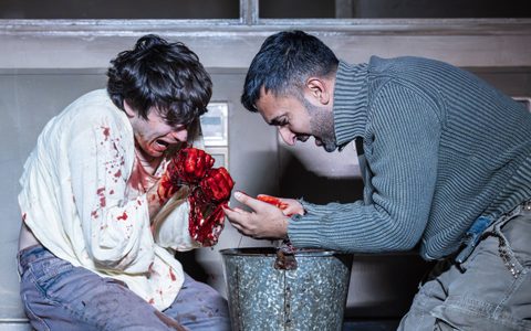Luke Cinque-White and Parth Thakerar kneel with a bucket between them. Luke is anguished and he looks down to both of his hands, which are covered in blood.
