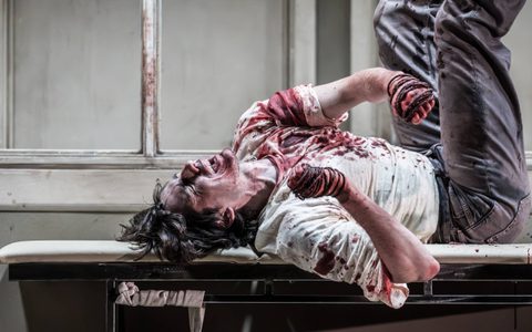 Luke Cinque-White lies on a cream doctor's bed, he is covered in blood and there is rope wrapped around each of his hands. His face is anguished and his eyes are closed.