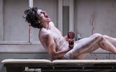 Luke Cinque-White screams on a cream doctor's bed. His hands are wrapped in rope and he wears no clothes.