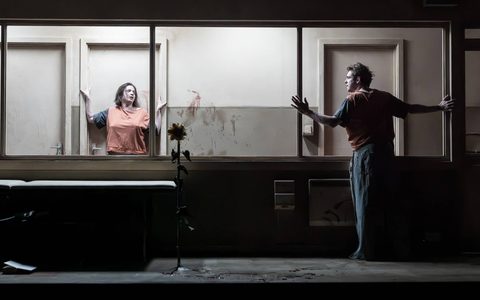 Jack Riddiford stands pressed up a glass window in a dark room. Pearl Chanda stands on the other side pressed against the back wall. Red blood is smeared against the glass window.