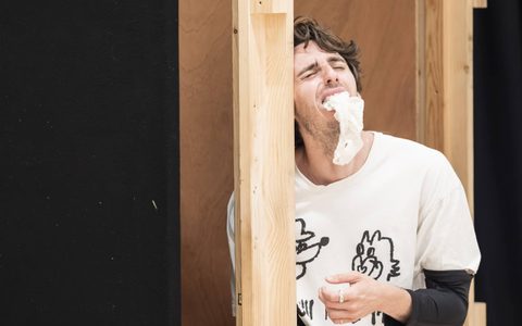 Luke Cinque-White stands against a wooden frame. He has a white bandage in his mouth and is wearing a pained expression on his face.