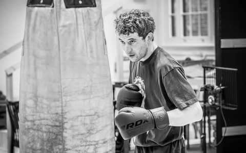 Josh O'Connor stands in front of a large punch bag wearing large black boxing gloves.