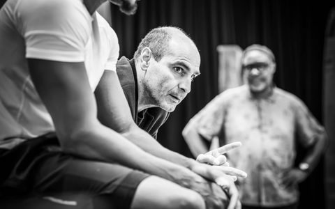 Zubin Varla sits with his arms rested forward on his kneed. Hi looks to the side with a serious expression and points forward.