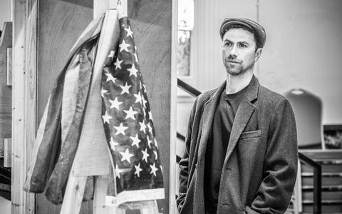 Richard Fleeshman stands wearing a long coat and a flat cap. He has both hands in his coat pockets and wears a serious expression.