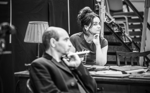 Zubin Varla and Hayley Squires sit at a wooden desk. They both rest their left hand against their chin.