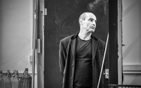 Zubin Varla stands in a long black coat with his hands behind his back. He looks to the left hand side with a focussed expression on his face.