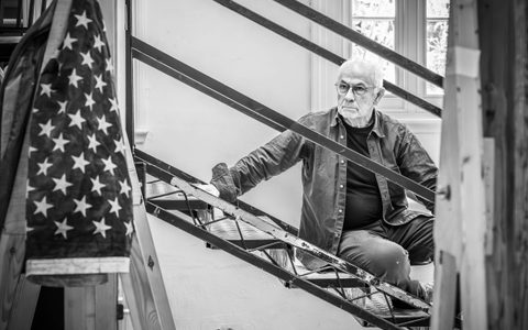 Nicholas Woodeson sits on a staircase leaning on step with one hand. He has a serious expression on his face and is looking forward.