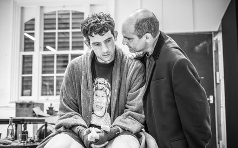 Josh O'Connor sits on a table wearing a towel robe and fingerless gloves. Zubin Varla stands next to him talking in his ear.
